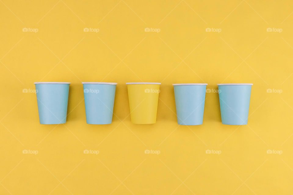 Four blue and one yellow cardboard glasses lie in a row in the center on a yellow background, fly lay close-up. The concept of easter, spring, minimalism, yellow and blue.