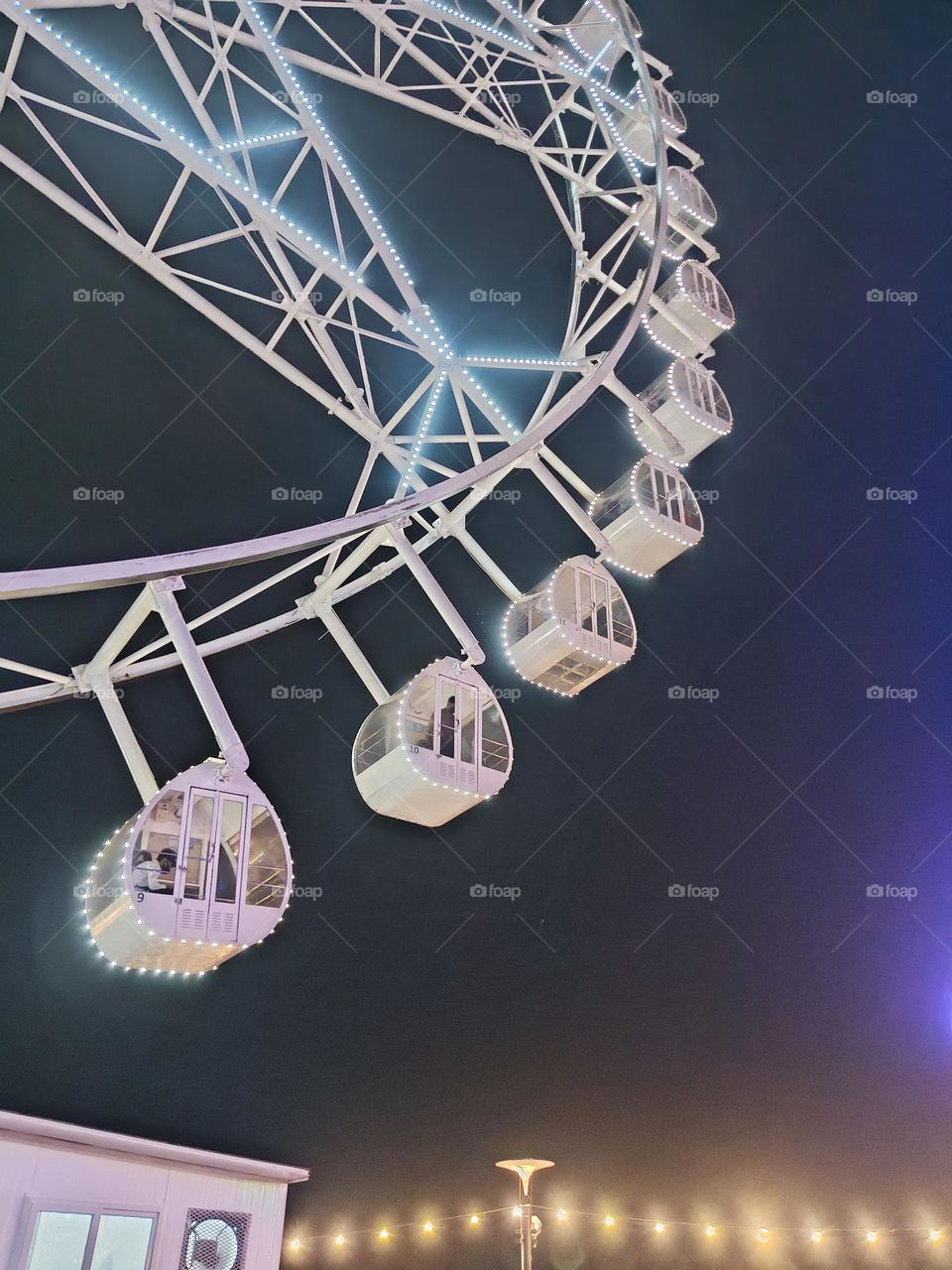 Romantic Perris wheel with lovers inside. It has white geometric figures with dark background. The lights give it a modern and classy look. The cabins are pear shaped.