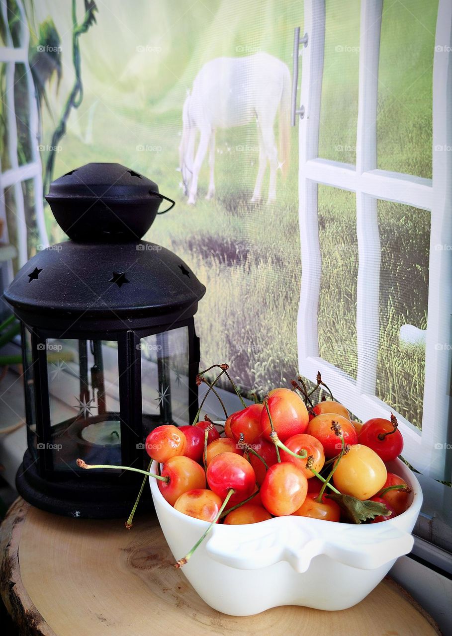 On the windowsill of the window lies a wooden cut of wood, on which stands a white cup with cherries and a black lantern.  The windows are open.  There is a net on the window, behind which a green meadow with a white horse