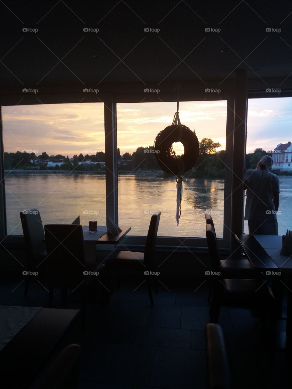 From a restaurant hall, a photo of an Austrian river at the sunset with German confine on the other side of the river. In first plan tables and chairs for customers.