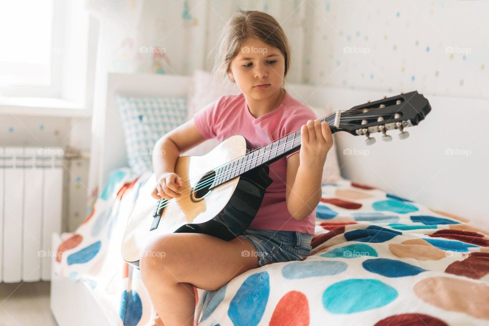 Cute tween girl in pink t-shirt play guitar sit on bed in bright room at home