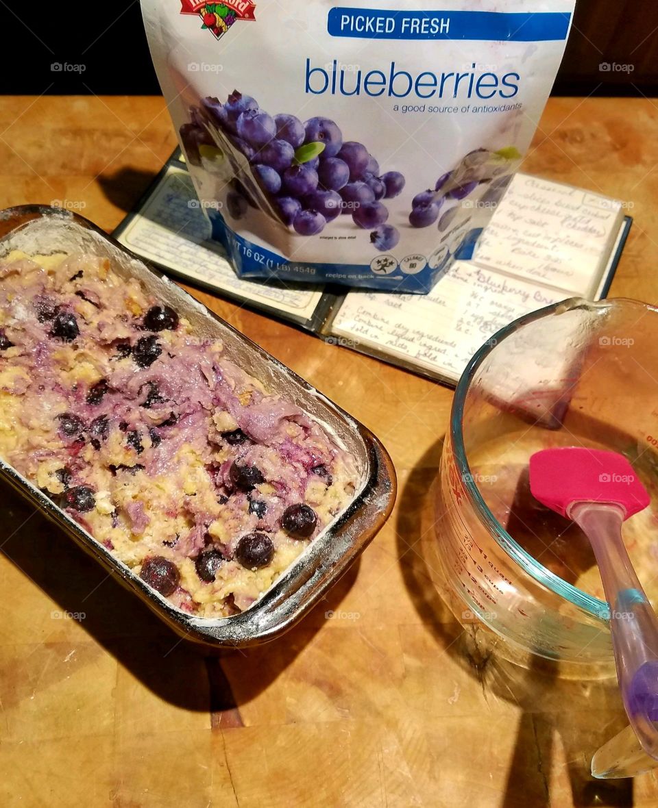 Blueberry bread in baking dish ready to go into hot oven. Recipe book & utensils are nearby.