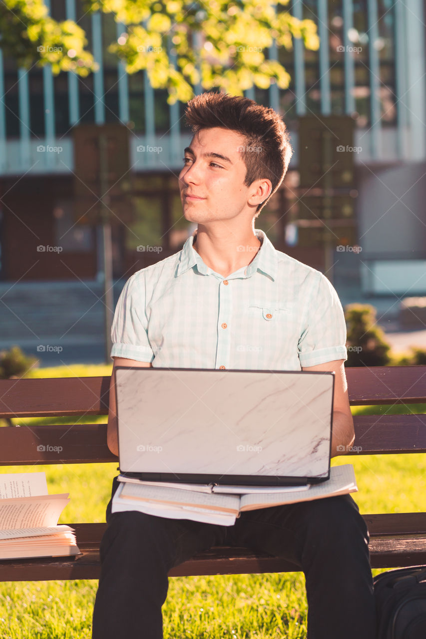 Student working on a laptop using books and notes sitting on a bench in a park. Young boy wearing a blue shirt and dark jeans