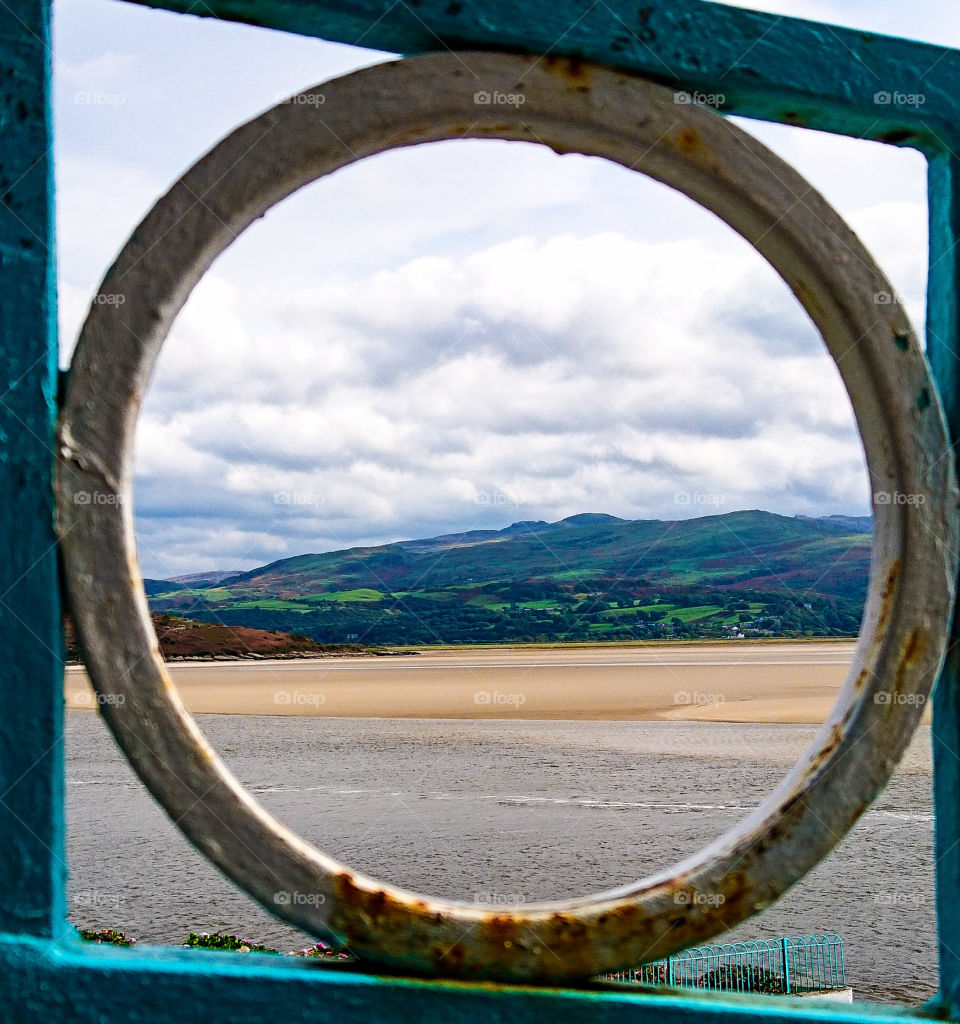 beach and countryside view at Port meirion in Wales. Greens, browns, blues and golden. Through a circular decorative railing.