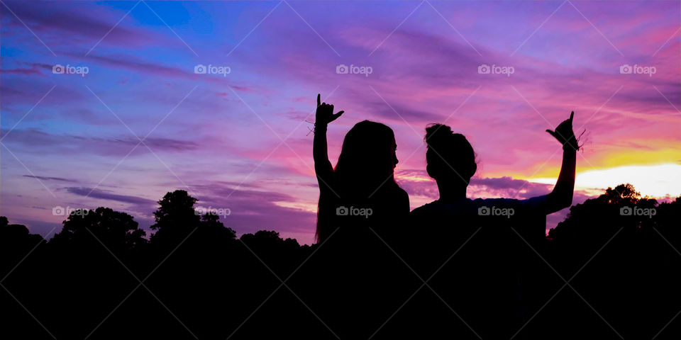 Two silhouetted girls throw up hang ten hand signs against a beautiful cotton candy colored sunset.