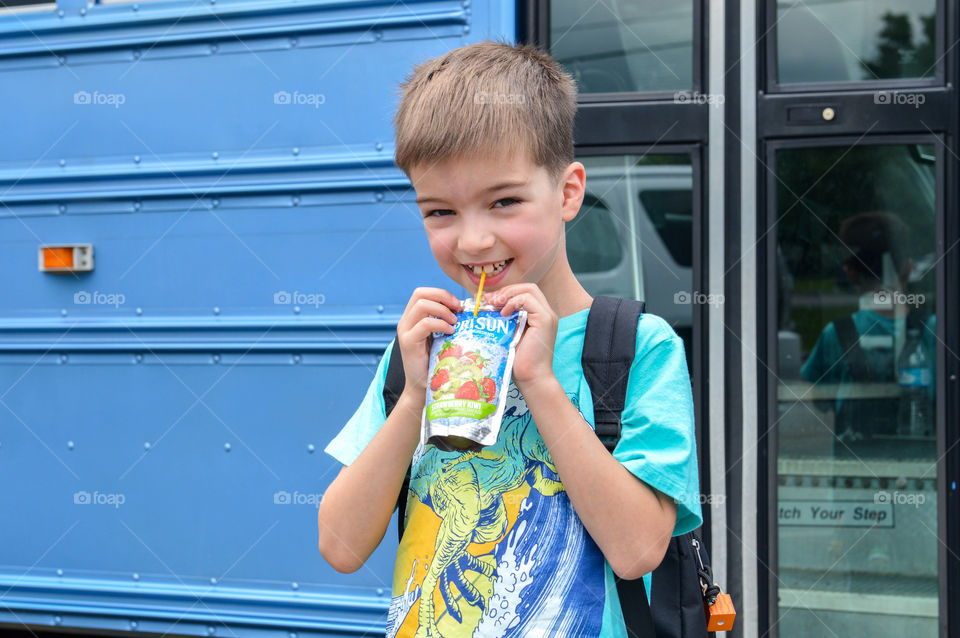 Young boy smiling and drinking from a drink pouch in front of a blue school bus