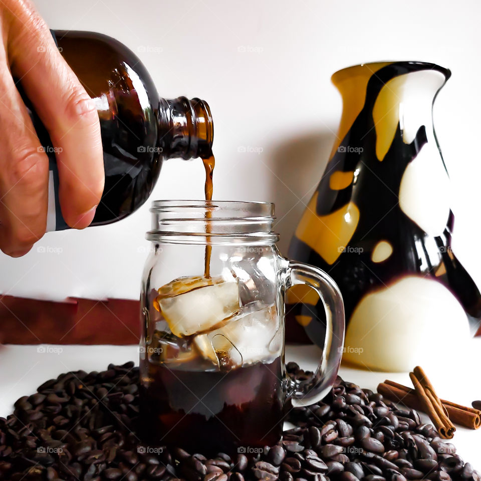 Pouring cold brew black coffee in iced filled mason jar with handle, surrounded by whole coffee beans.