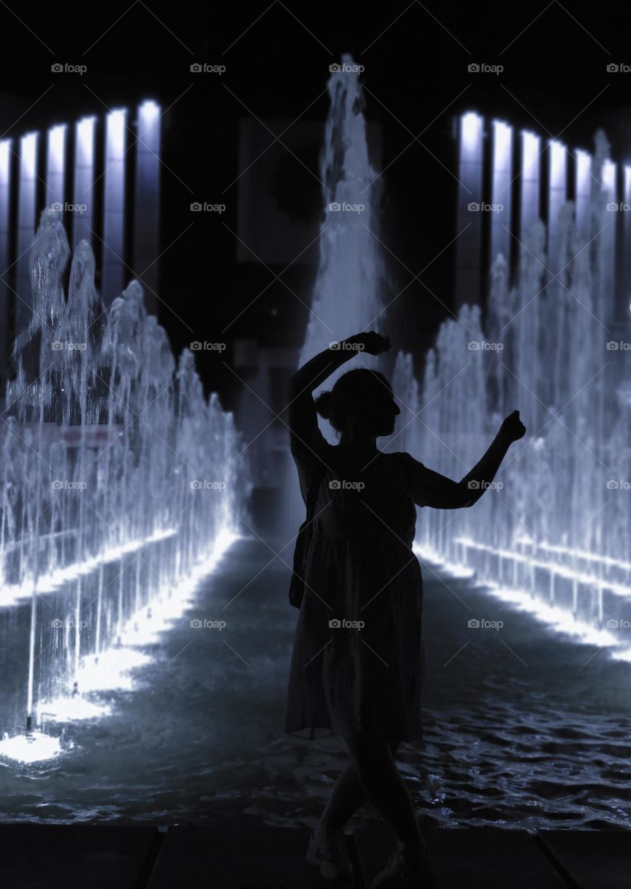 A shadow of a woman dancing in front of a fountain in the darkness