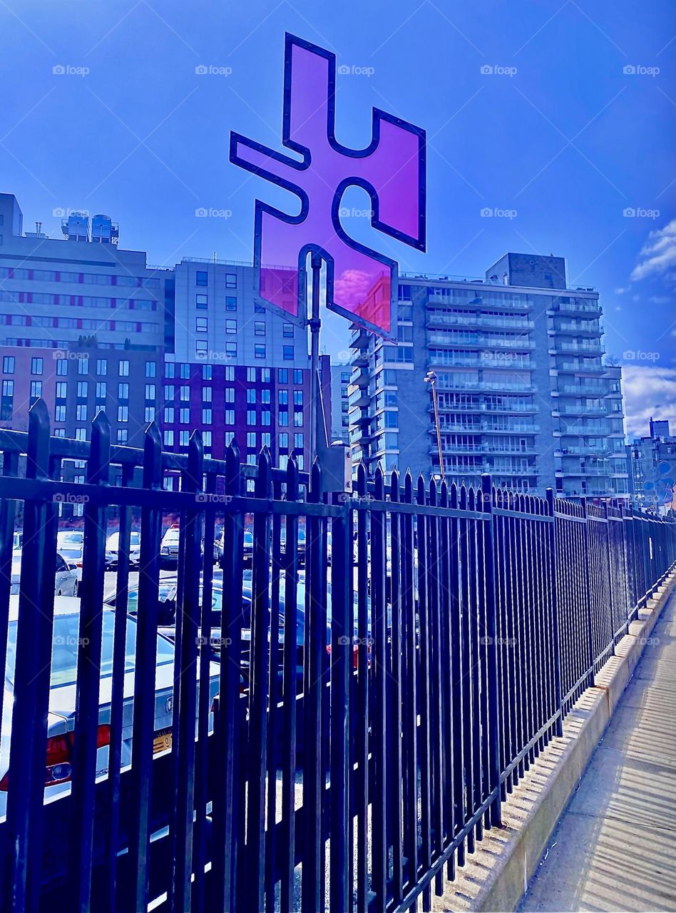 Another geometric shape reminding of a jigsaw puzzle piece attached to a fence in LIC, Queens. The fence w multiple parallel metal poles blends in nicely w the shapes of high rise buildings with multiple rows of windows. 2022. Hypnotic Productions