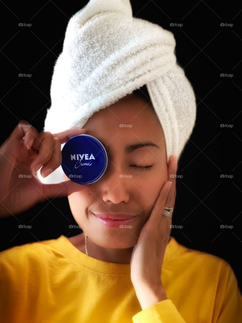 A woman holding a Nivea creme with white wrapped towel in her head and hand on her checks. Portrait in black background. 