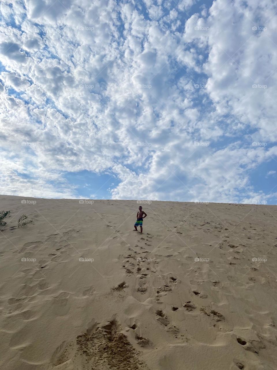 This image shows a person standing on a sand dune under a sky filled with scattered clouds. The footprints in the sand suggest a climb, and the scene gives a sense of vast openness and adventure.