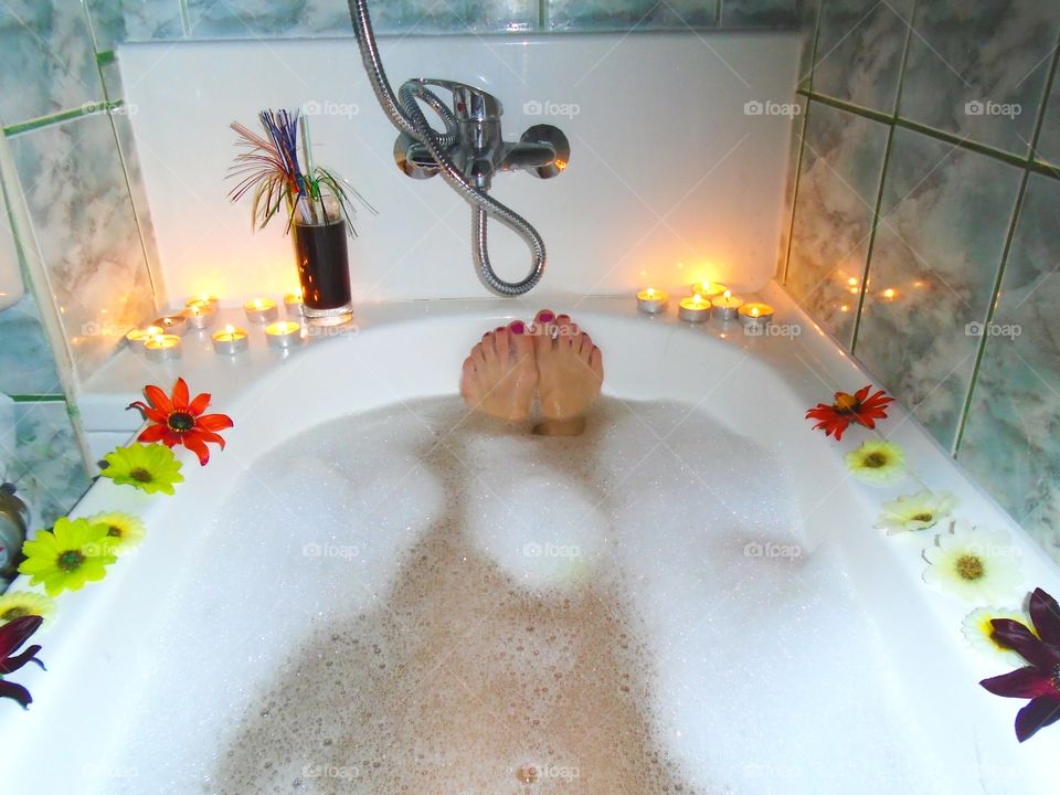 Woman's feet with red nail polish in bubble bath with candles and flowers