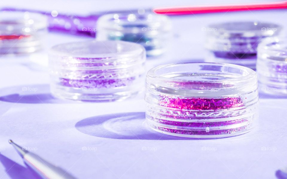 A set of clear little jars of glittery nail shadows on a lilac background, side view. Concept cosmetics and beauty salons.