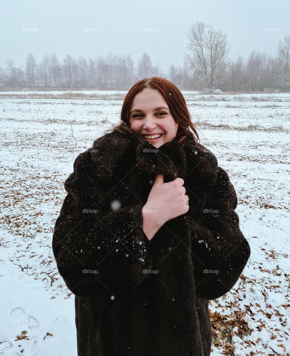 Winter portrait of a fair-skinned girl in a fur coat smiling at the camera and enjoying snowflakes in the middle of a wild field