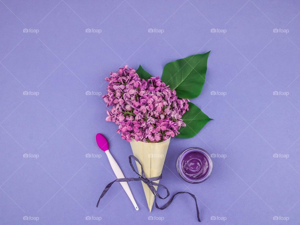 One branch of lilac in a wooden cone with a jar of cream and a brush for applying cream lie on a lavender background, flat lay close-up. The concept of natural cosmetics.