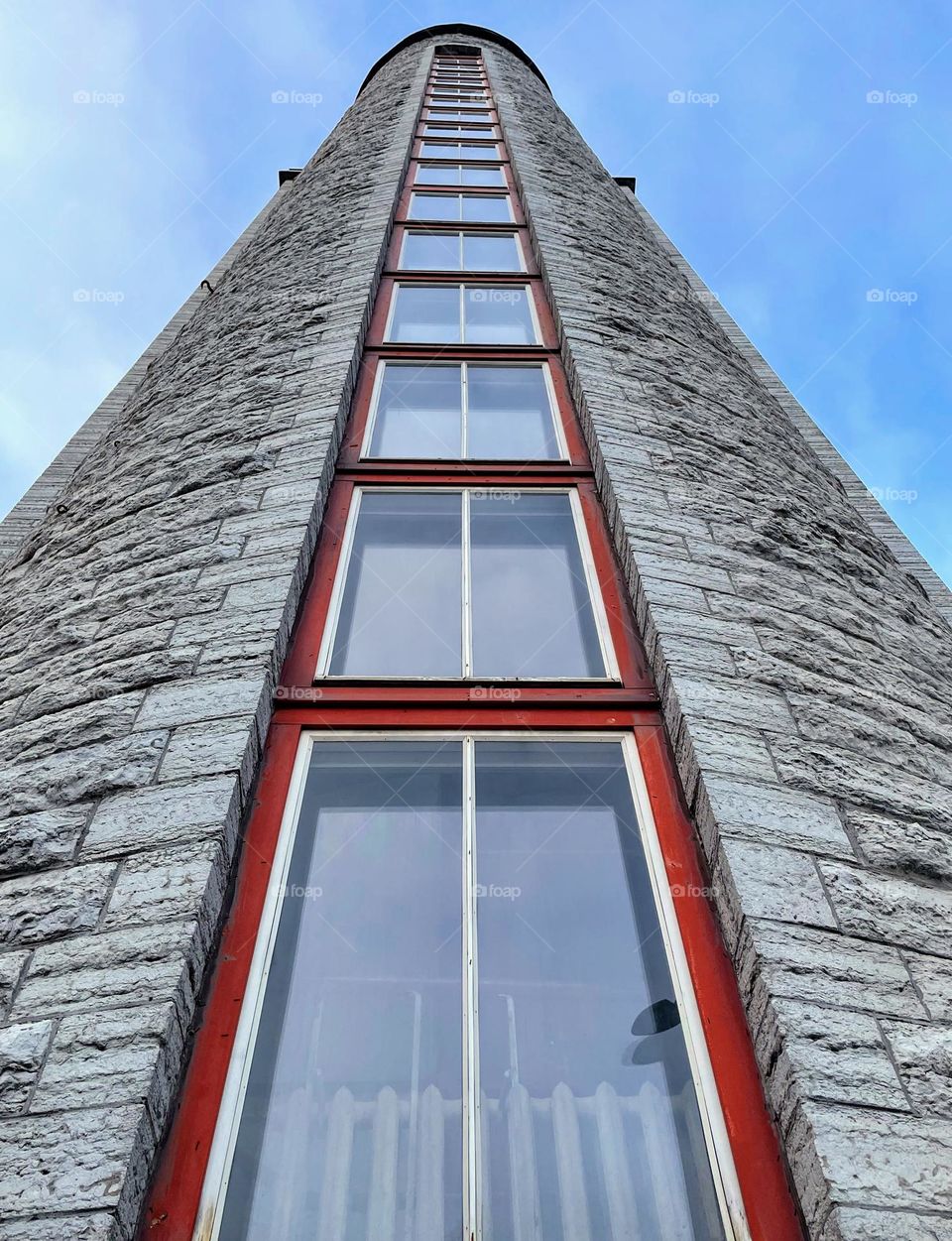 Bottom up view of high narrow round rough stone building with big windows with red frames 