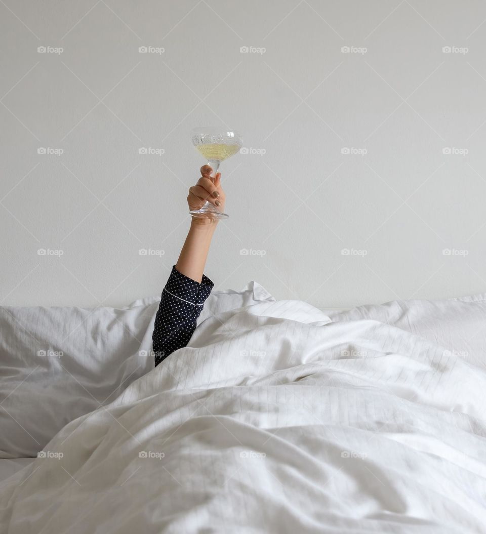 Person in bed, covered with white linen, holding glass of wine, champagne.