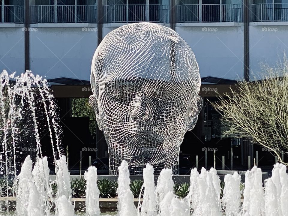 A mesh face sculpture faces the fountain in the city