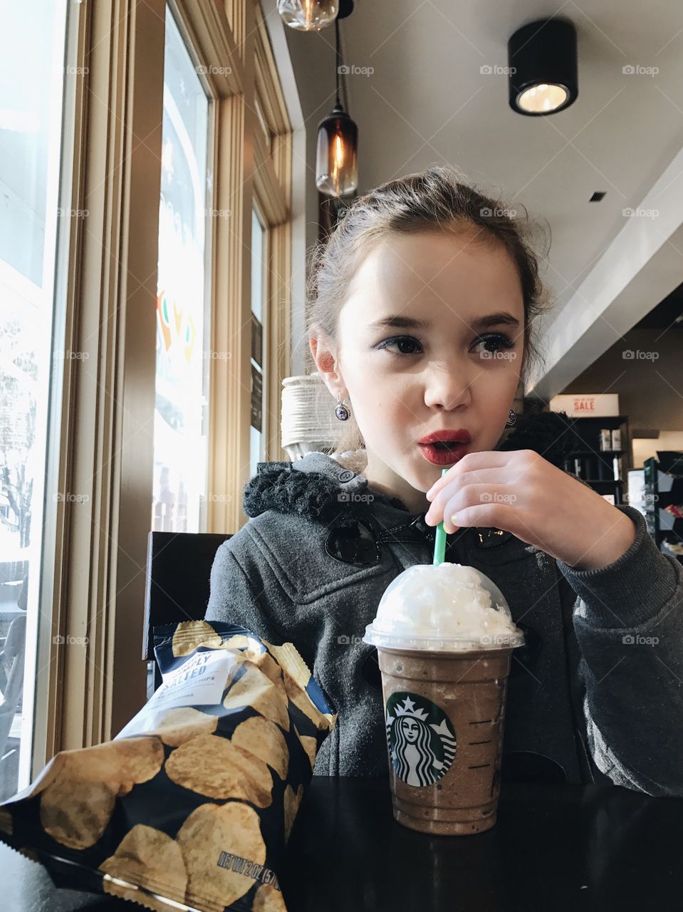 Cute, little girl with lipstick on drinking a Starbucks Frappuccino in the winter with a bag of chips and her coat on. 