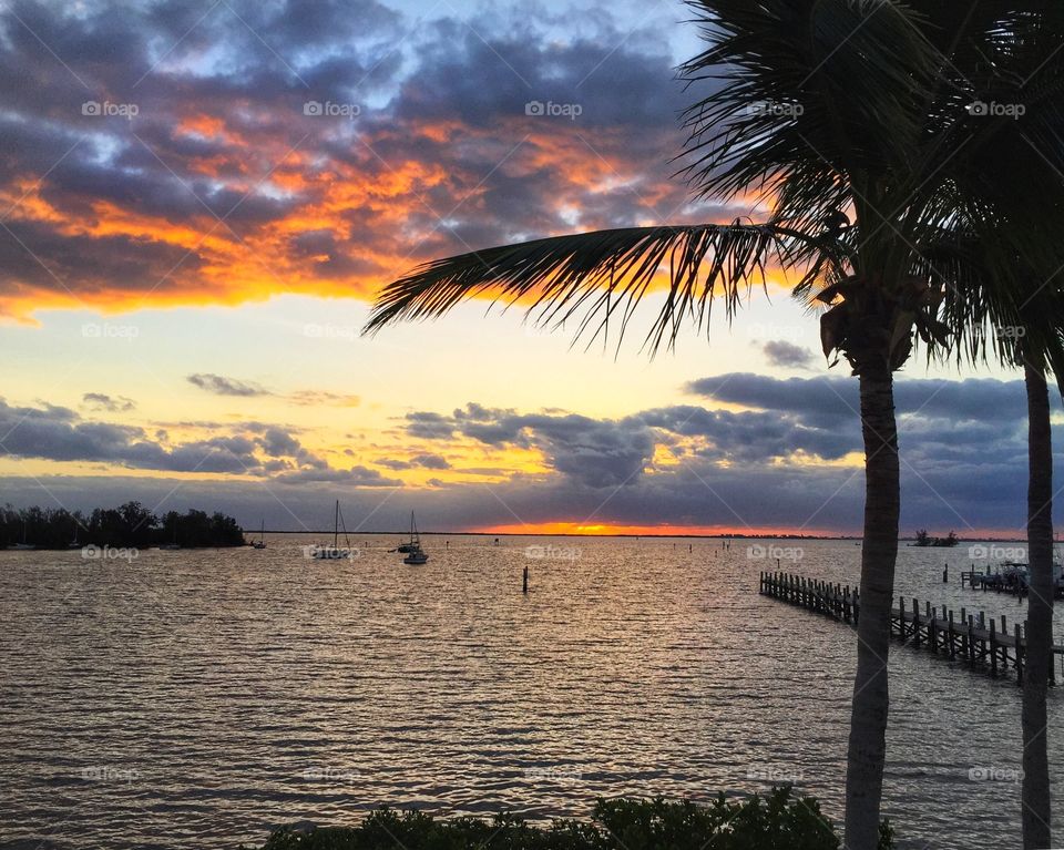 Colors of the sunrise reflected in the clouds above and on the river below with silhouettes of palm trees, anchored boats and a dock.