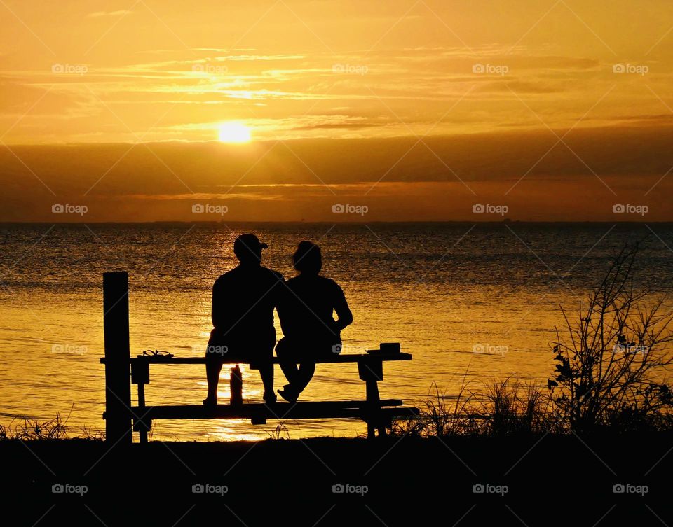 Life is good. A romantic couple sitting on a picnic table spend time together alone, away from family and friends watching a magnificent sunset descending over the shimmering bay