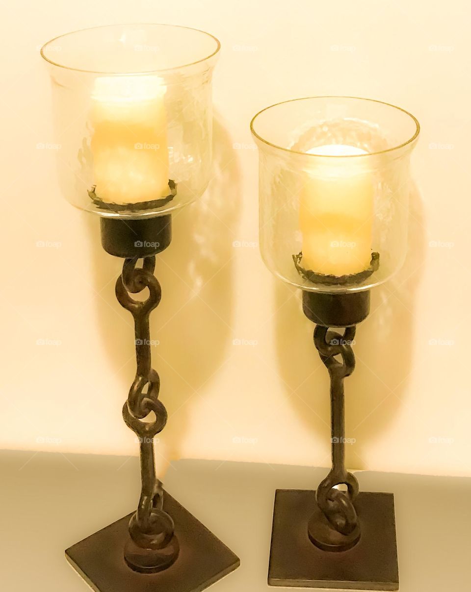 Two rustic candle holders on a table with yellow/white candles casting shadows on the wall. 