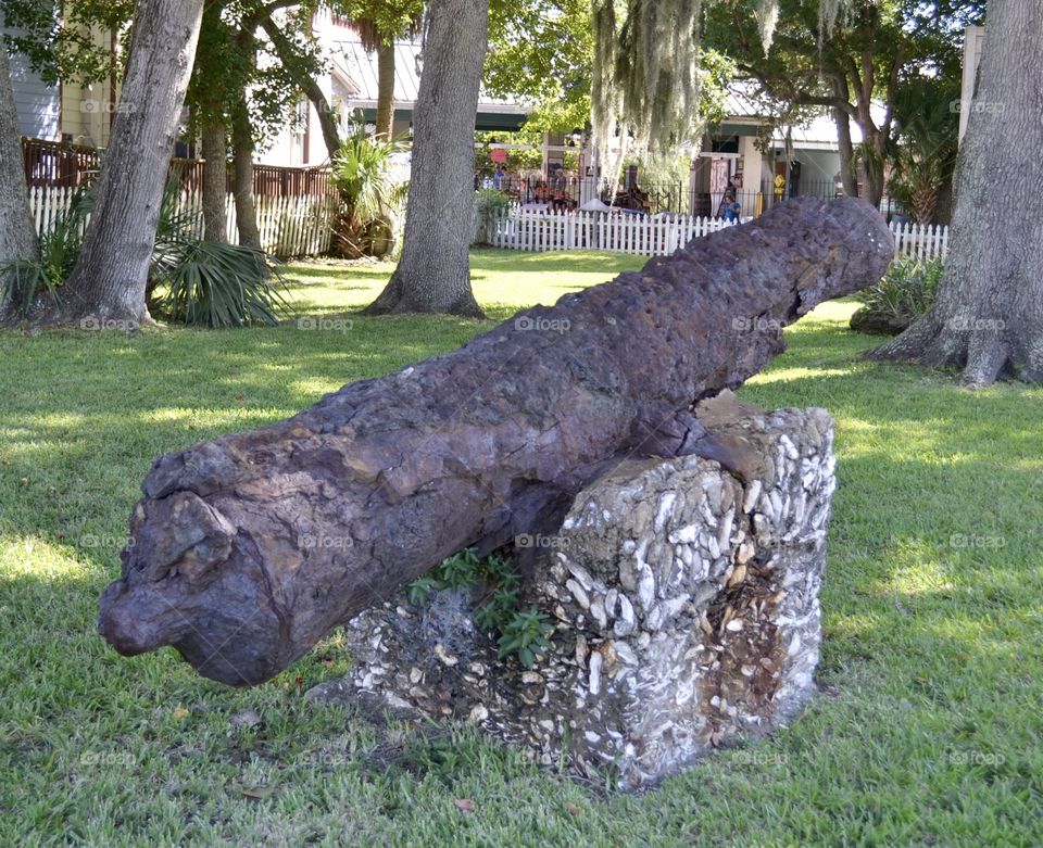 A very old rusty metal cannon resting on a block of coquina rock on green grass with palm trees nearby 