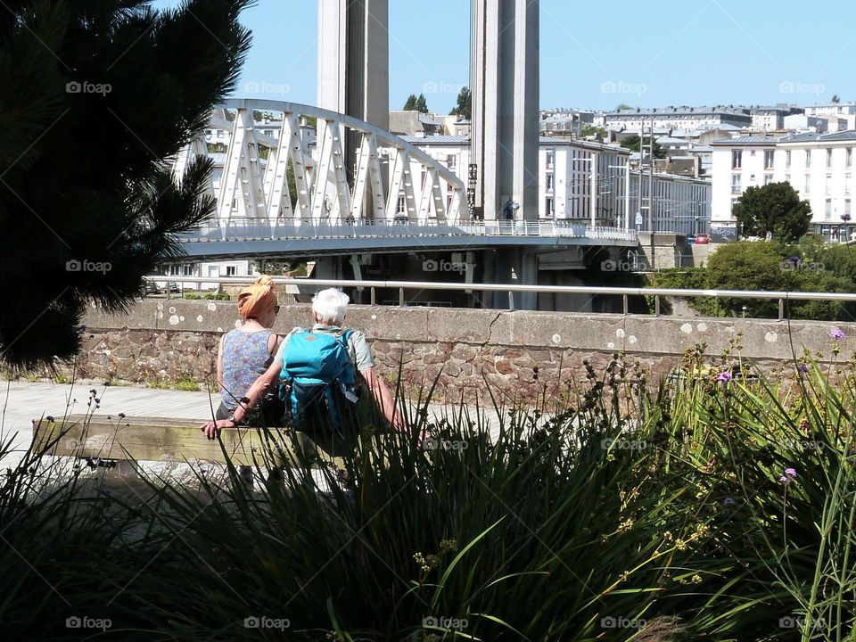 Two tourists, a senior couple, are sightseeing in town, the man is carrying a hiking backpack on his back. They are seated on a bench facing a bridge.