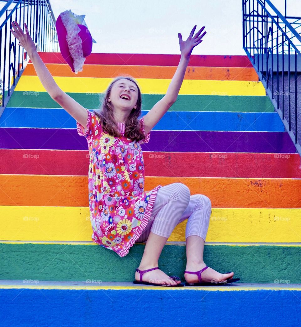 Joyful girl on rainbow-coloured stairs tosses her hat 