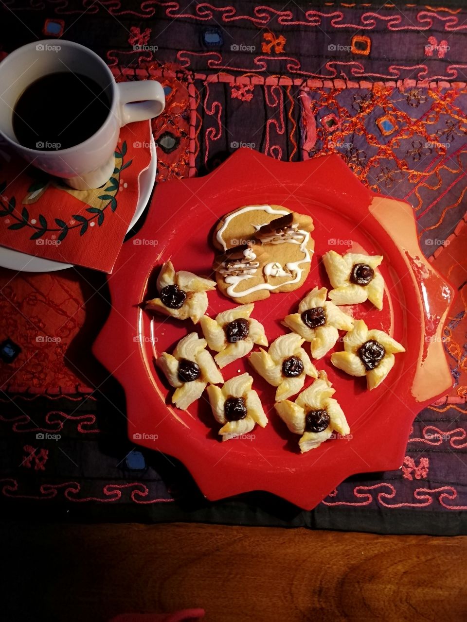 A star shaped red plate full of small pastries with plum jam. A gingerbread decoration with two sugary coated trees with golden pieces. A coffee cup on the multicoloured napkin on a white plate.