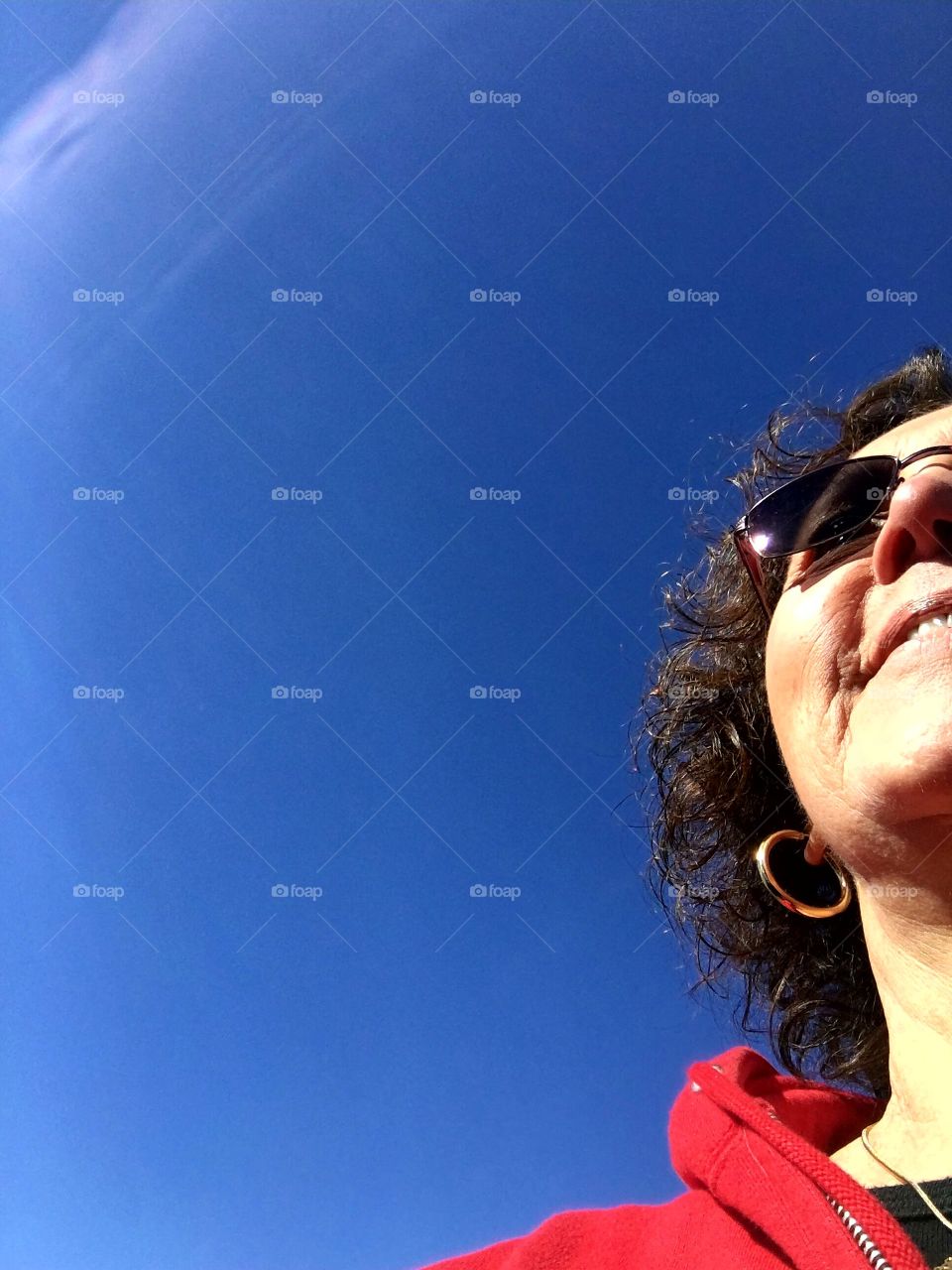 Smiling womans face with pic taken from below her. Happy girl outside walking on chilly day. Wearing sunglasses, sweatshirt & a smile with her gold hoop earring showing. Sky is blue with no clouds.