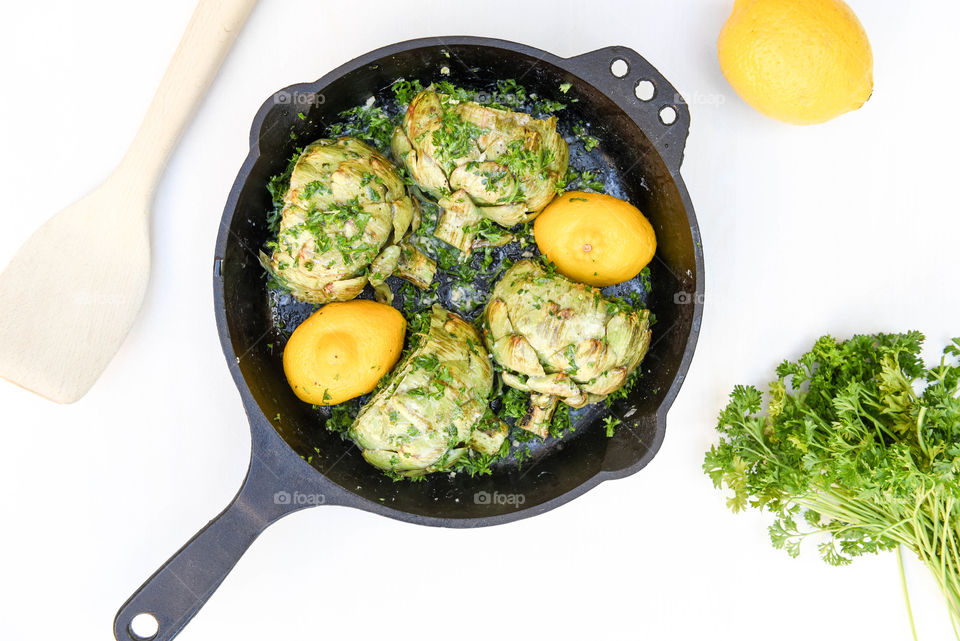 Flat lay of roasted artichokes with lemon in a cast iron skillet surrounded by a wooden spoon and parsley on a white background