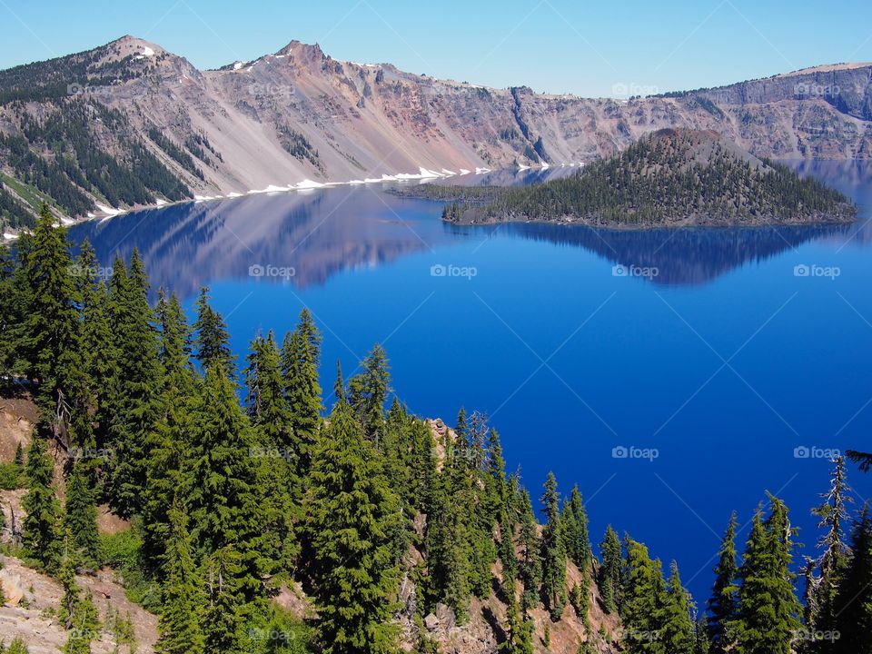 The jagged rim reflecting into the rich blue waters of Crater Lake in Southern Oregon on a beautiful summer morning with perfect clear blue skies. 
