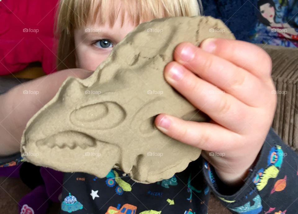 Close up photo of a male child holding a sand sculptured skull of a triceratops dinosaur. One eye visible. 