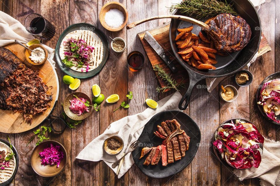 "Top view of a large food feast.
Next to a large cast iron skillet with meat and potatoes is a small bowl with sliced ​​limes, tortillas, and cabbage."