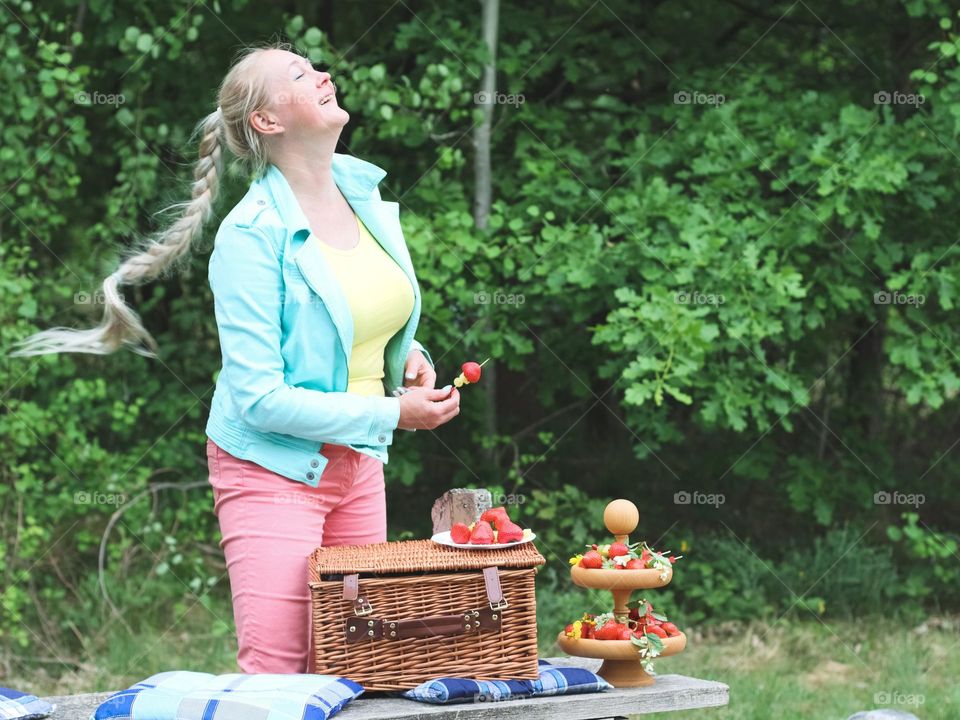 One beautiful young caucasian blonde girl having fun shaking her head and long braided hair while standing by a wooden bench with a wicker basket and fruits in the forest at a picnic on a summer day, side view close-up. Concept happy people, picnic t