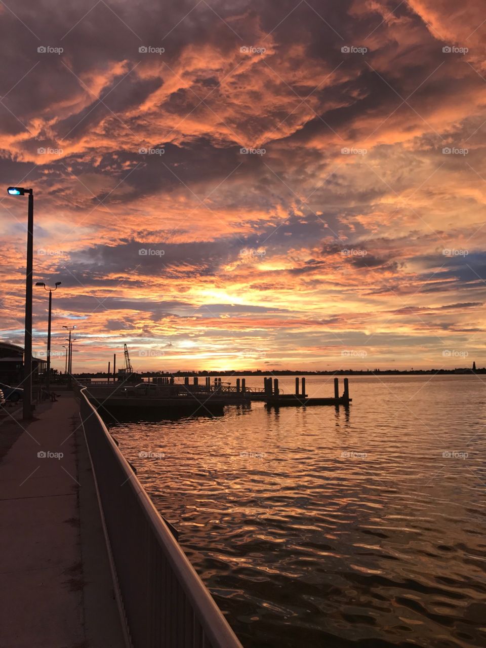 Sunset over the water along the bridge, the sun illuminating the clouds in shades of yellow, orange and purple.
