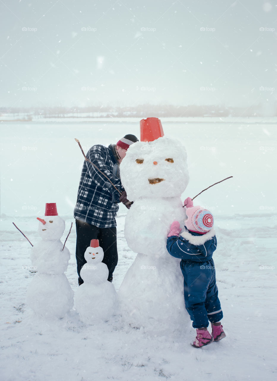 Man and his little daughter making a snowman, spending time together outdoors, having fun on snow in wintertime. Dad and kid wearing warm clothes