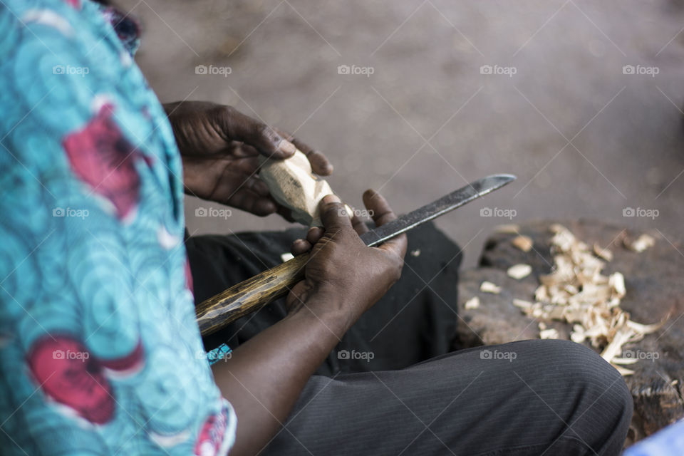 An African American man carved a piece of wood with a knife carefully as he makes traditional wooden sculptures by hand with some wood shavings on the side as his favorite hobby
