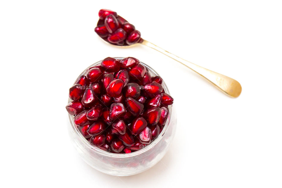 Closeup of red pomegranate seeds in a glass with single white spoon on a white background. Love this delicious red summer fruit. Encourages a healthy diet and lifestyle.