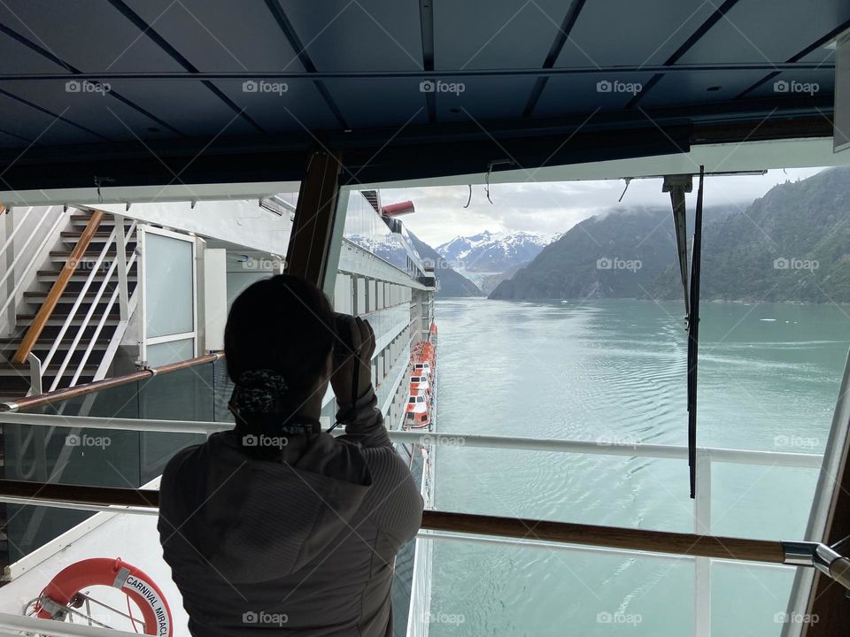 A woman with binoculars looking out at a glacier from a ship’s bridge while sailing in Alaska.