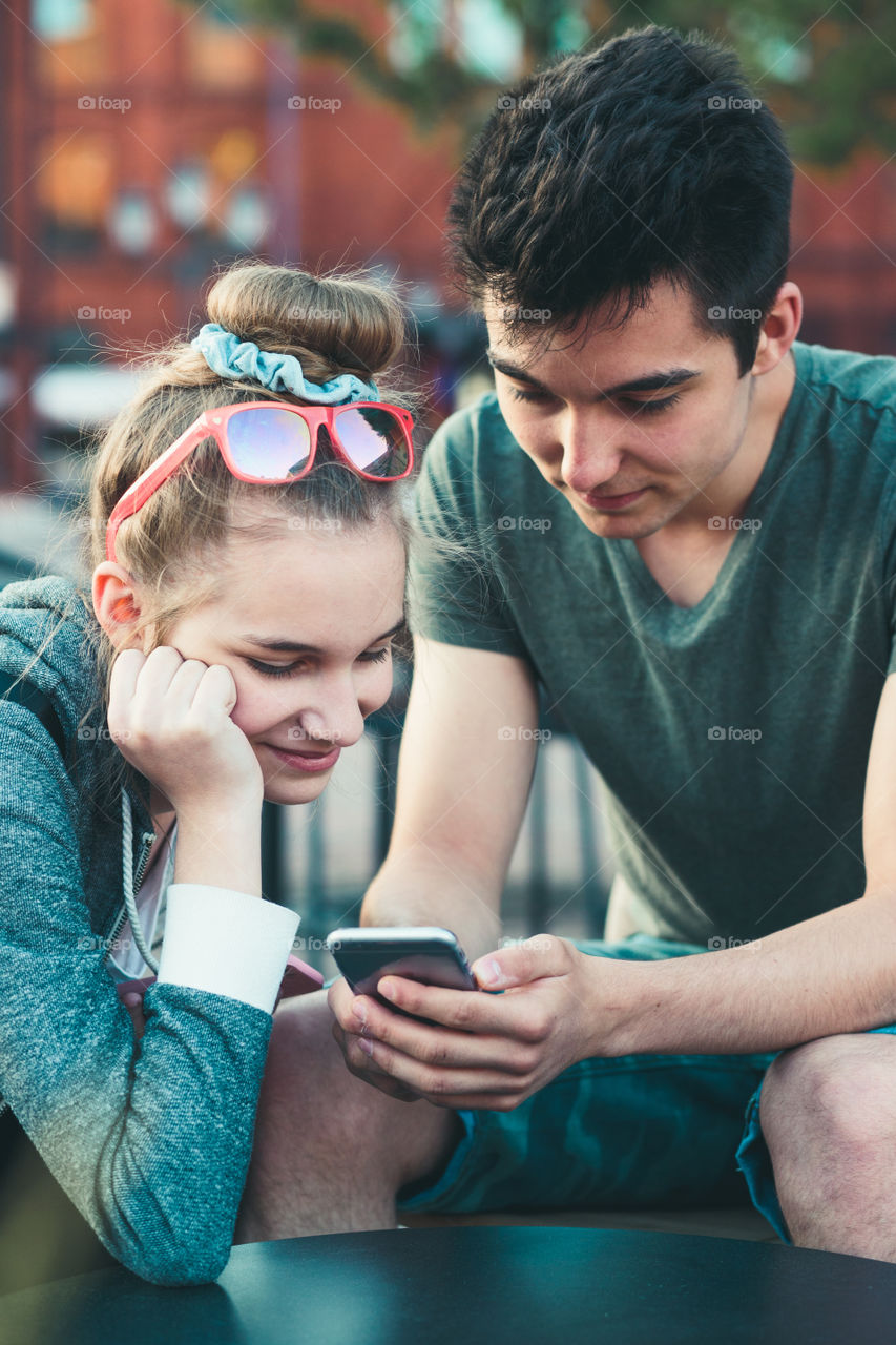 Couple of friends, teenage girl and boy, having fun with smartphones, sitting in center of town, spending time together