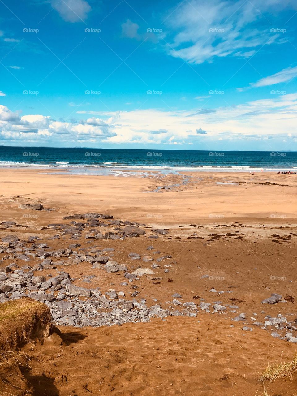 Fanore Beach glows under a light blue sky with white puffy clouds, meeting the deep blue Atlantic. The shore shifts from pale brown to rich orange-brown sand, scattered with rocks in a mix of earthy tones.