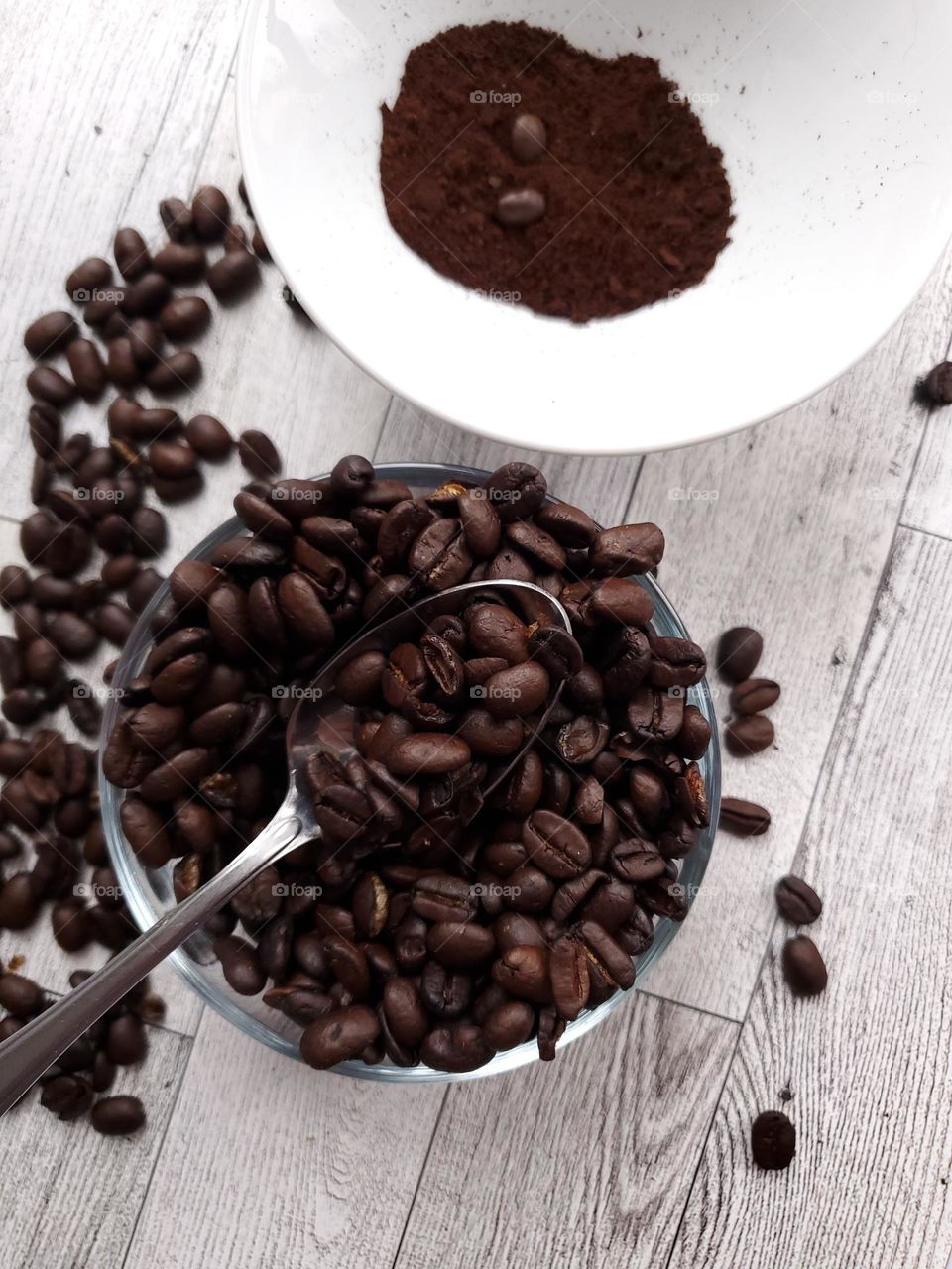 A clear glass bowl with coffee beans and a spoon.  A white bowl above with freshly grounded coffee beans. Loose coffee beans on the surrounding light colored diagnol wood surface.