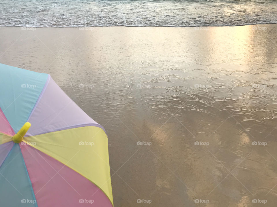 part of colorful pastel umbrella on clear sand beach with sea wave and glowing sunset light reflects on  the sand