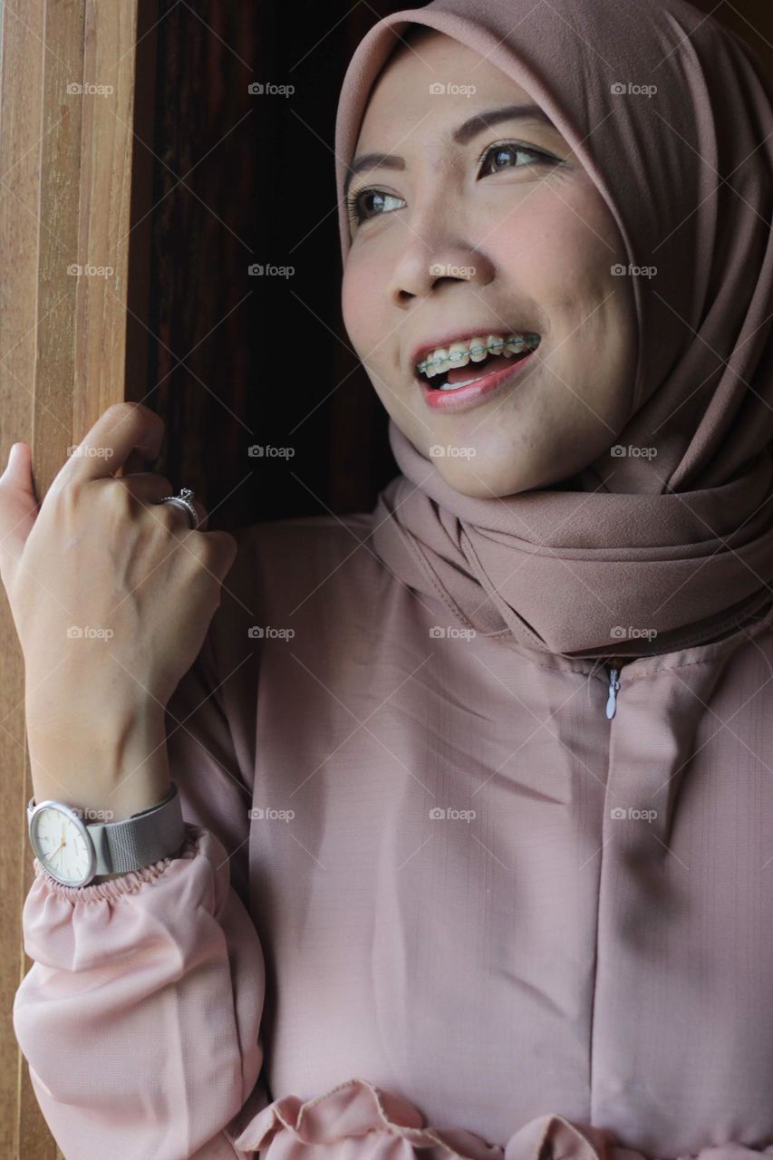 Portrait of a young Asian Muslim woman wearing a pink (Chewing Gum color) hijab, smiling out a window.