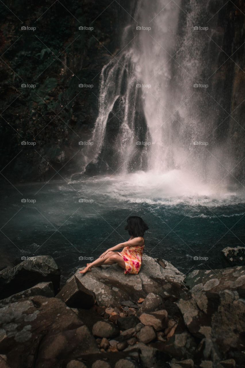 a woman sitting on a rock in front of a waterfall that has green water