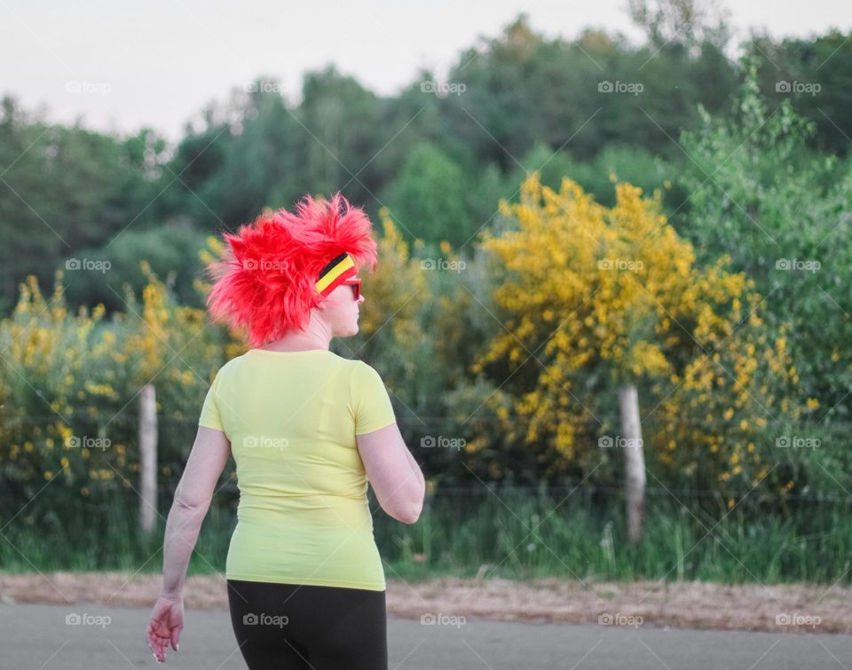 Portrait of a caucasian middle-aged woman in a yellow t-shirt, black leggings and a red wig from the back walks down the street towards a field in belgium, close-up side view. Belgium day concept.