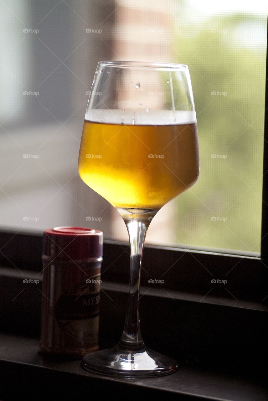 A glass of beer sits next to a bottle of cooking spice on a window ledge.