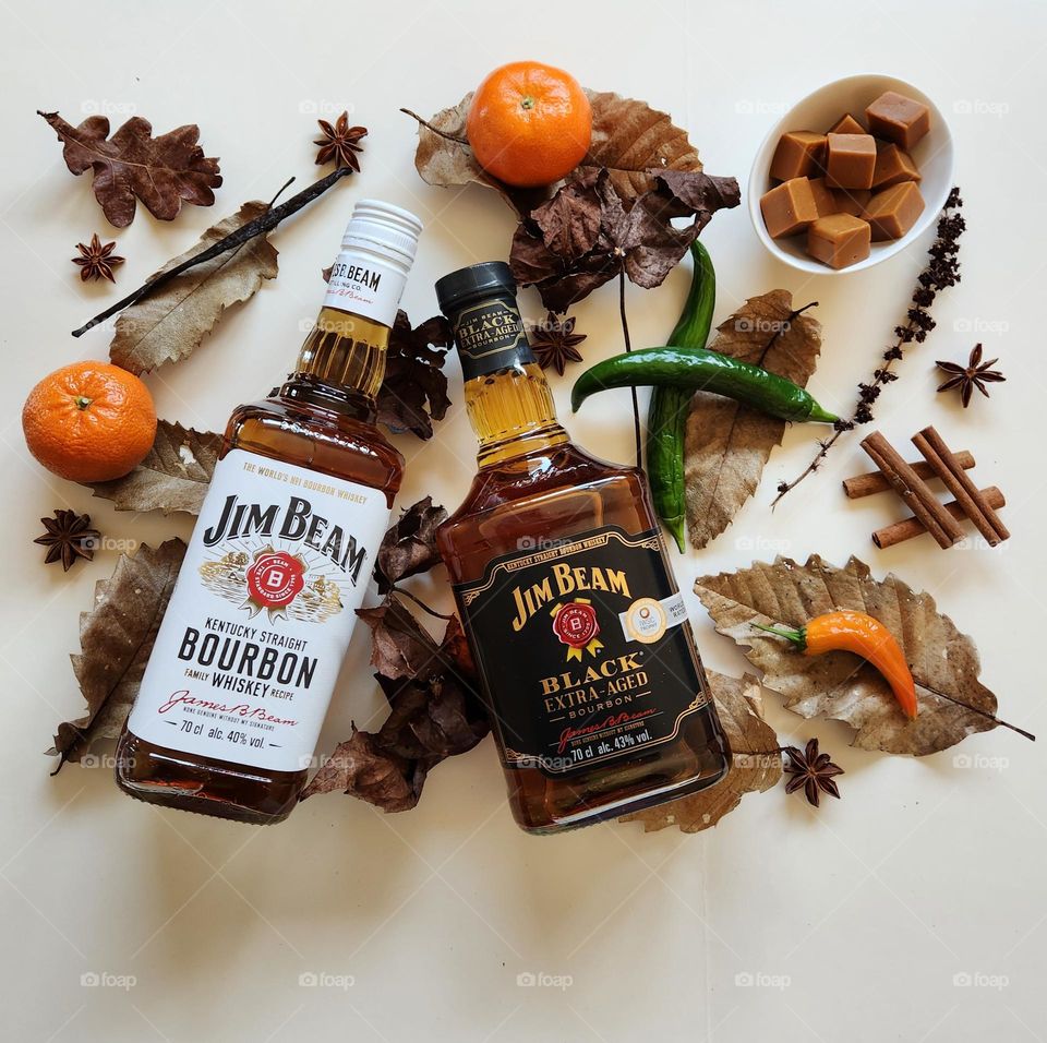 Winter Jim Beam Kentucky Straight & Black Extra Aged bourbon flatlay composition on cream surface and autumn leaves with fresh piments, cinnamom sticks, vanilla pod, mandarines and caramels in a china bone little bowl